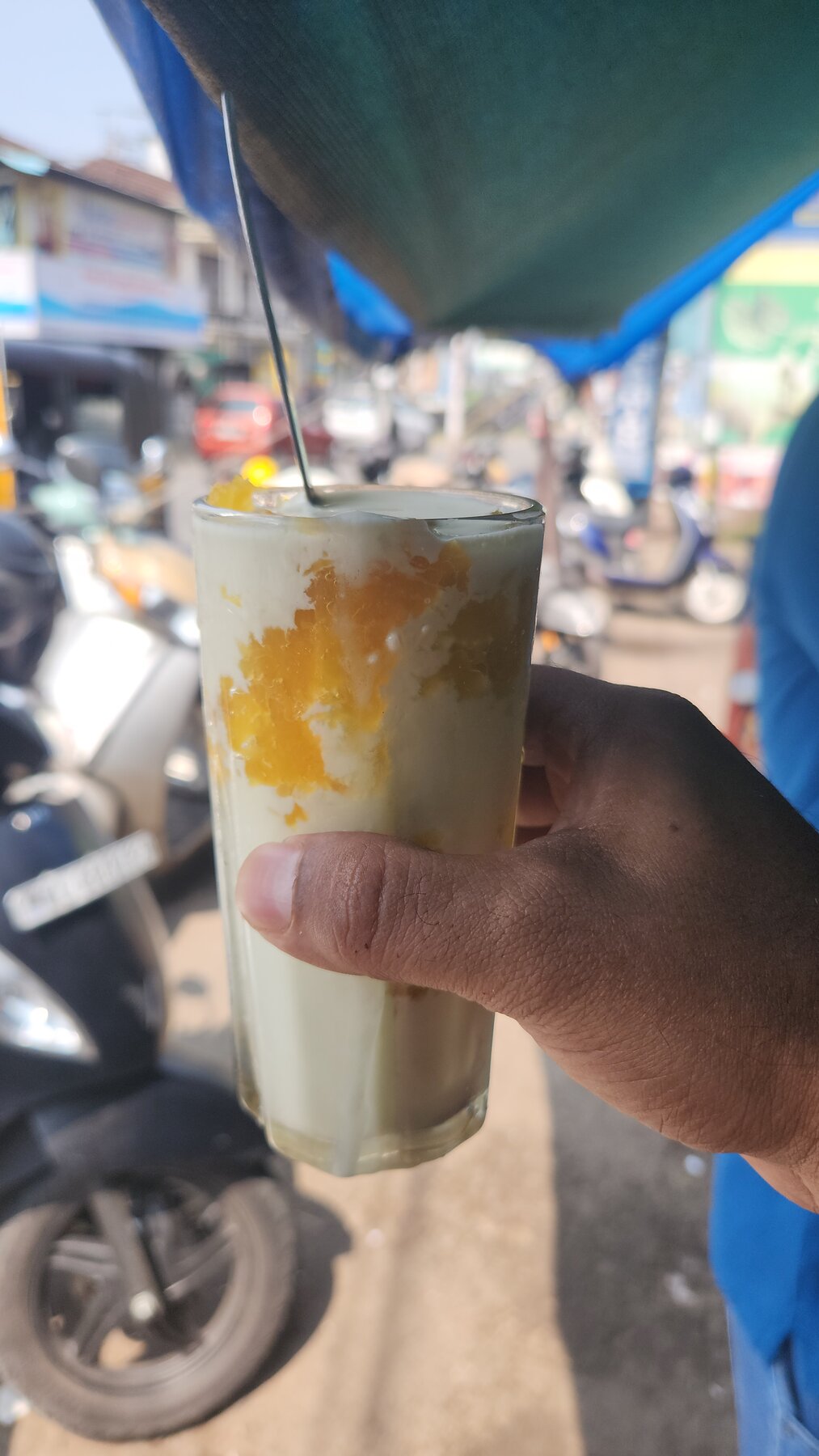 A hand holding a tall glass mango shake from Thomson Fruits with fresh mango chunks visible inside