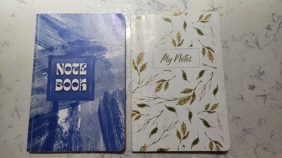 The two notebooks I bought my sons to track their investments — one blue with the word NOTEBOOK on the cover, one cream with a floral My Notes design