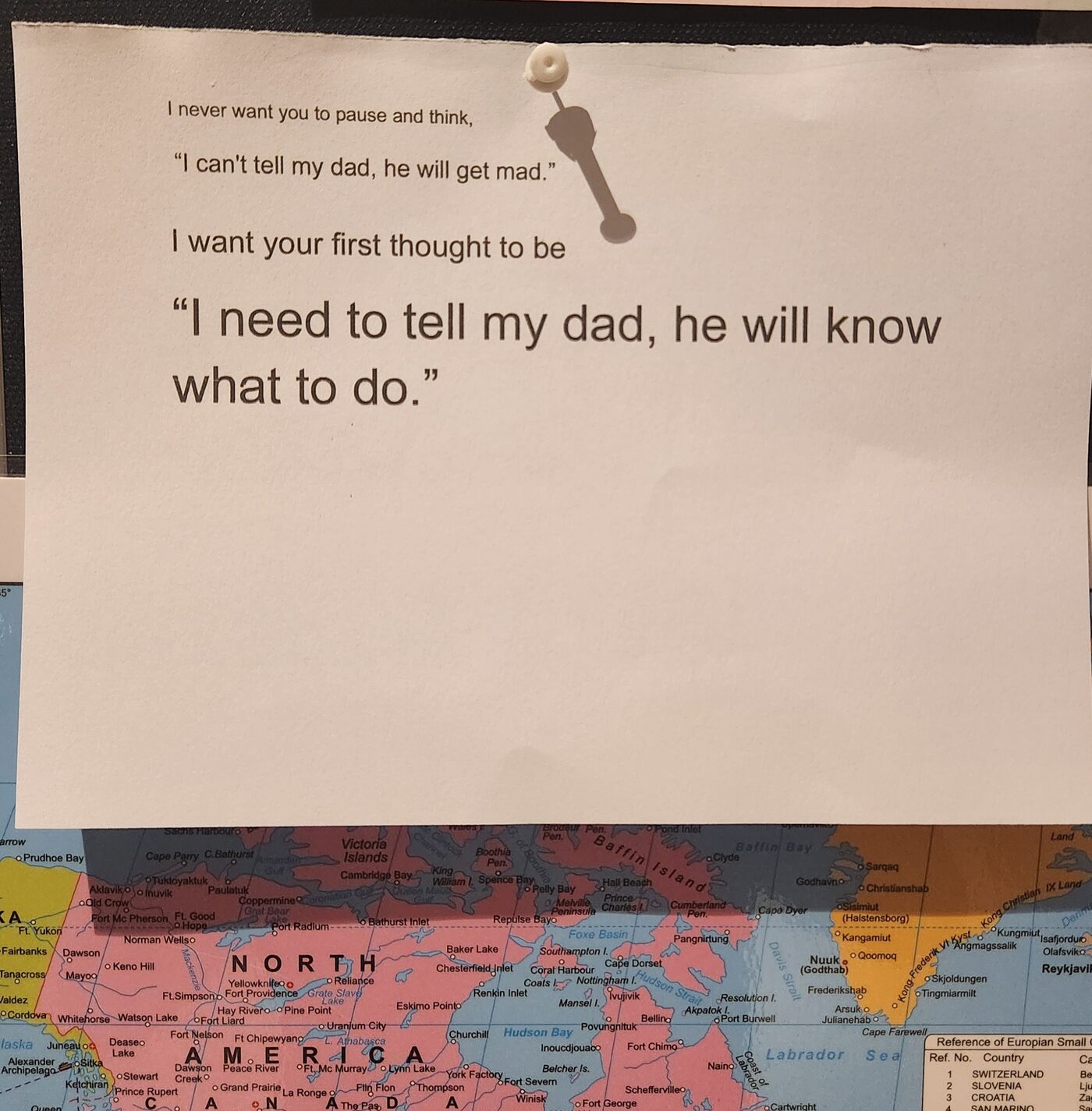 A printed note pinned to a board above a world map, reading: I need to tell my dad, he will know what to do